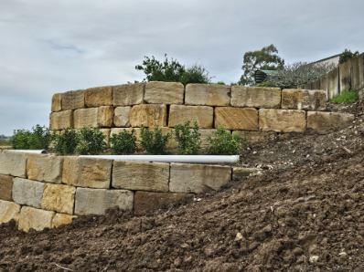 A finished sandstone retaining wall with a drainage pipe outlet and garden bed planted on top