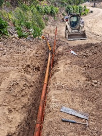 A drainage trench with orange pipe laid along its length, ready for backfill